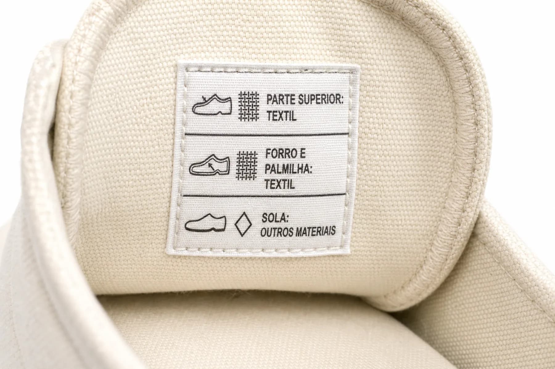 Macro photograph of a sewn-in material label on a canvas shoe reading parte superior têxtil, forro e palmilha têxtil, sola outros materiais